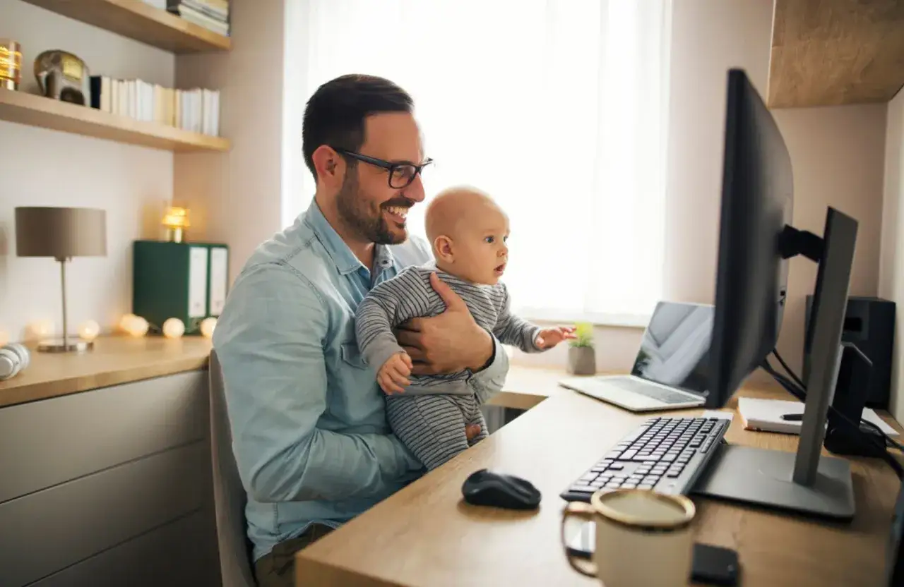 Un p&egrave;re souriant tient son b&eacute;b&eacute; tout en travaillant &agrave; son bureau. Il semble qu'il pr&eacute;pare sa lettre cong&eacute; paternit&eacute;.