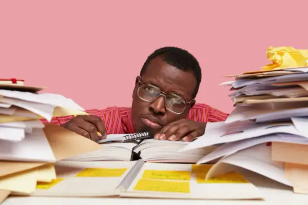 Un homme fatigué, entouré de piles de documents, semble dépassé par sa tâche. Il est peut-être en train de préparer un préavis foncia.