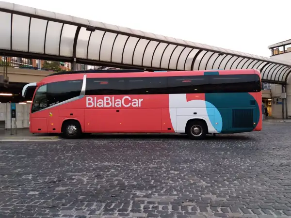 Un bus BlaBlaCar rouge et gris est garé sur une place pavée sous un auvent.
