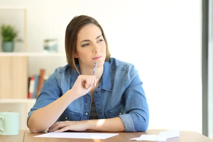 Femme songeant à une lettre de remise gracieuse impot taxe d'habitation, stylo à la main, devant des papiers.